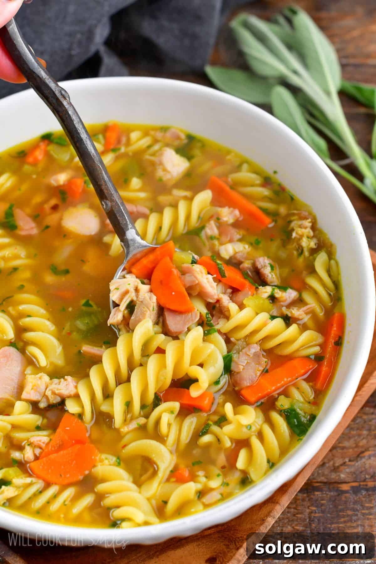 spooning out some comforting and rich turkey noodle soup from a white bowl.