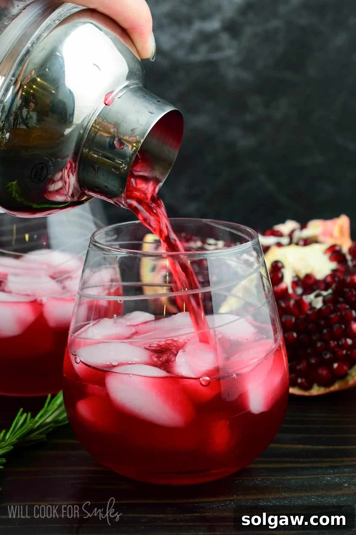 pouring the shaker gin cocktail into the clear glass next to rosemary sprig and open pomegranate.