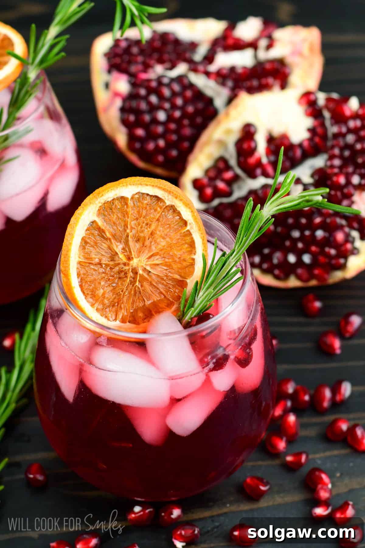 two glasses of pomegranate gin cocktail garnished with orange and rosemary and pomegranate seeds around.