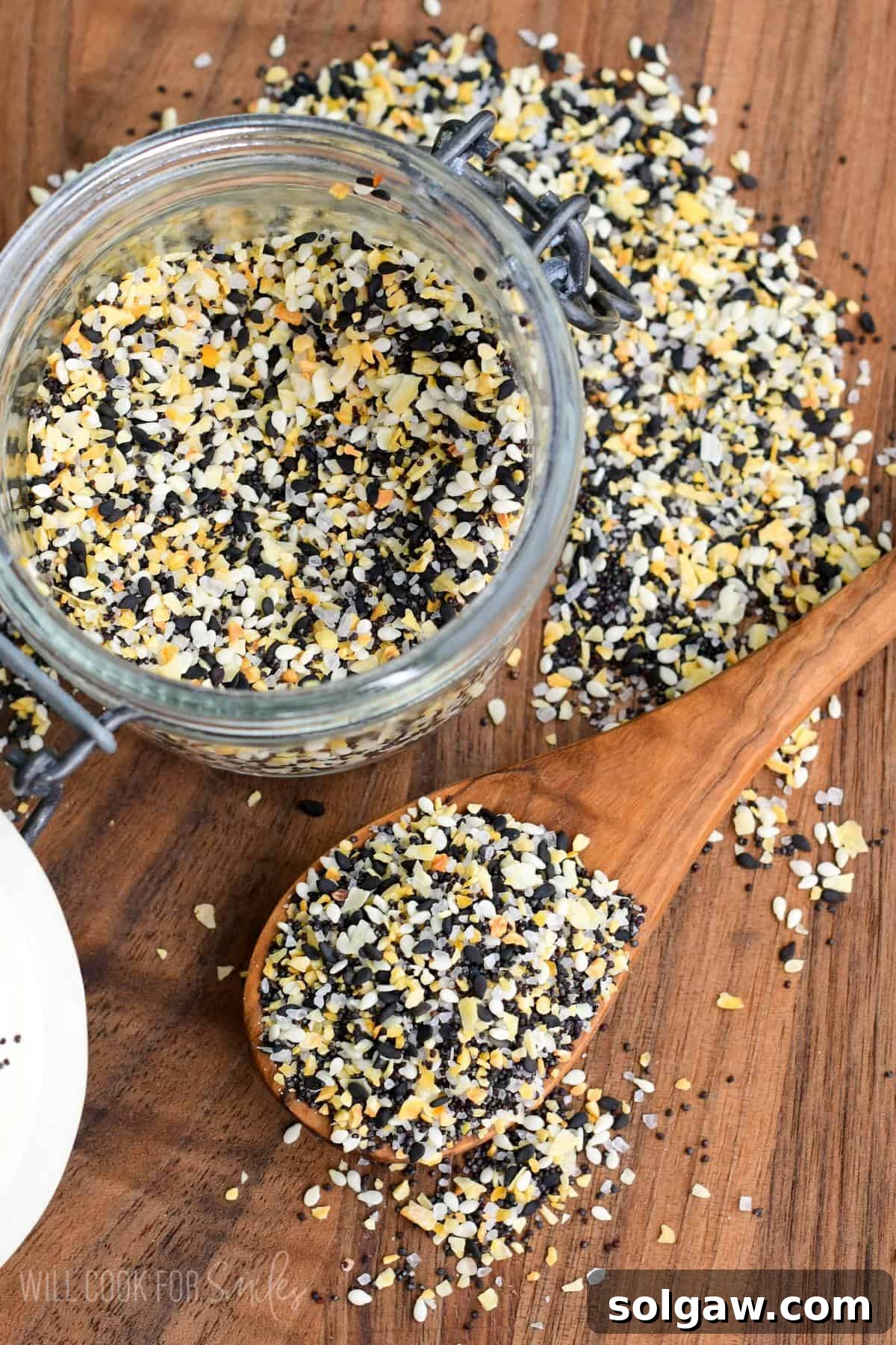 A jar overflowing with homemade Everything Bagel Seasoning, beside a spoon and some seasoning sprinkled on a wooden cutting board.