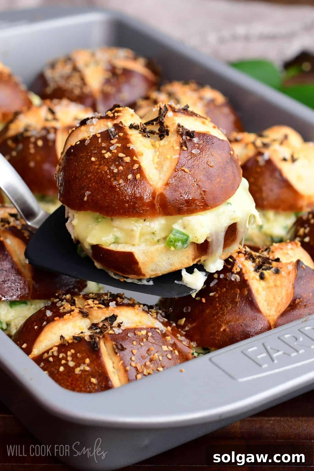 A baked jalapeño ranch chicken slider on a pretzel bun being lifted from the pan, showing melted cheese and creamy chicken filling. The golden-brown pretzel bun and oozing cheese promise a delightful bite.