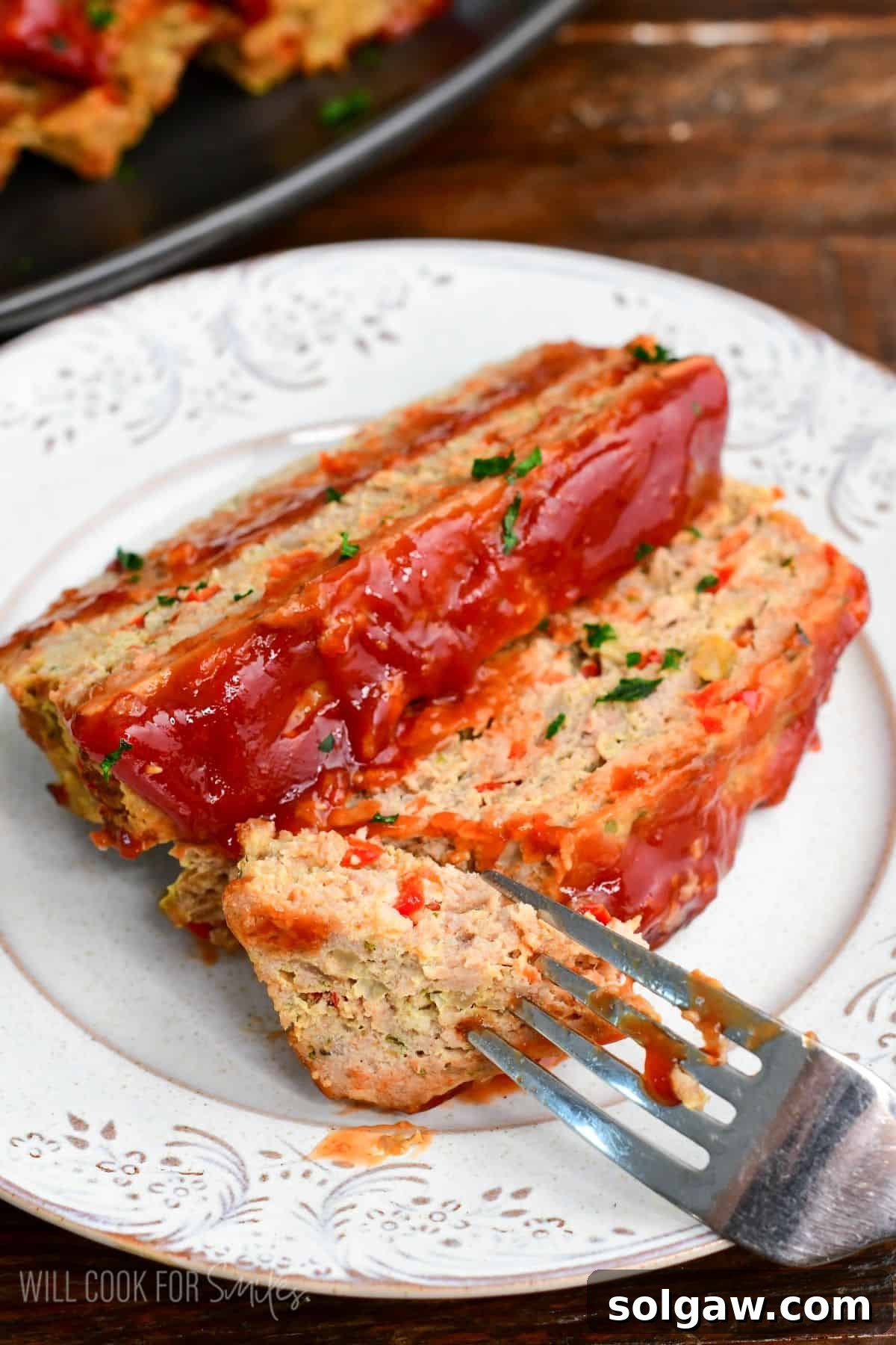 Two slices of turkey meatloaf on a plate with ketchup glaze and parsley, with a fork cutting into a tender bite.