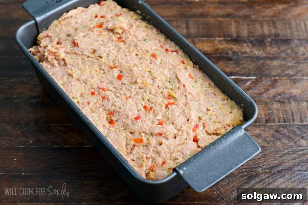 Uncooked turkey meatloaf mixture pressed evenly into a loaf pan, ready to bake, with visible diced red peppers and herbs throughout.