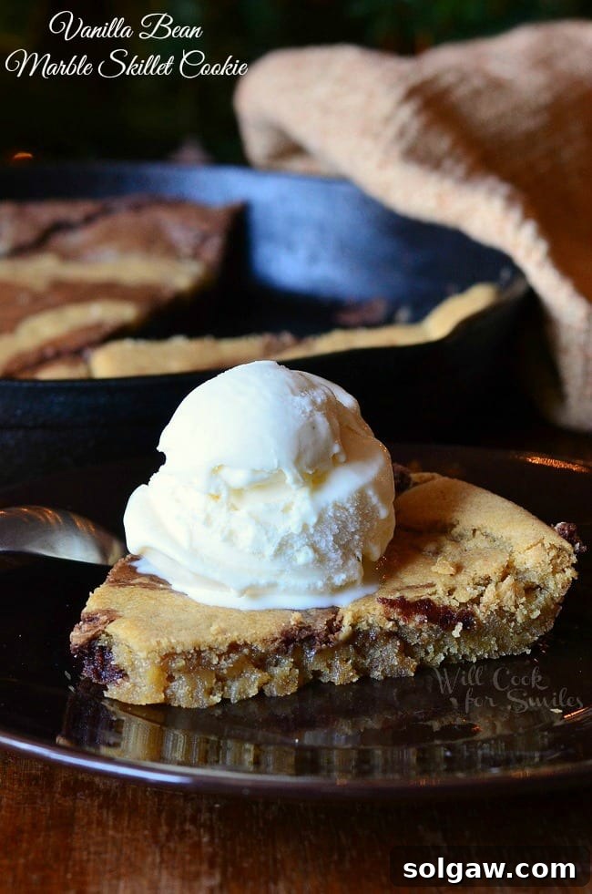 One slice of vanilla bean marble skillet cookie on a black plate, topped with a scoop of ice cream, positioned on a wooden table. The cast iron skillet with the remaining cookie is visible in the background.