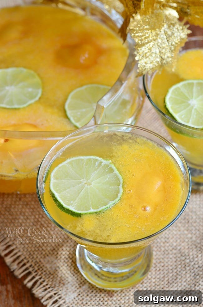 Champagne Sorbet Punch in a glass, with a full punch bowl in the background, adorned with lime slices. A perfect refreshing beverage for a gathering.