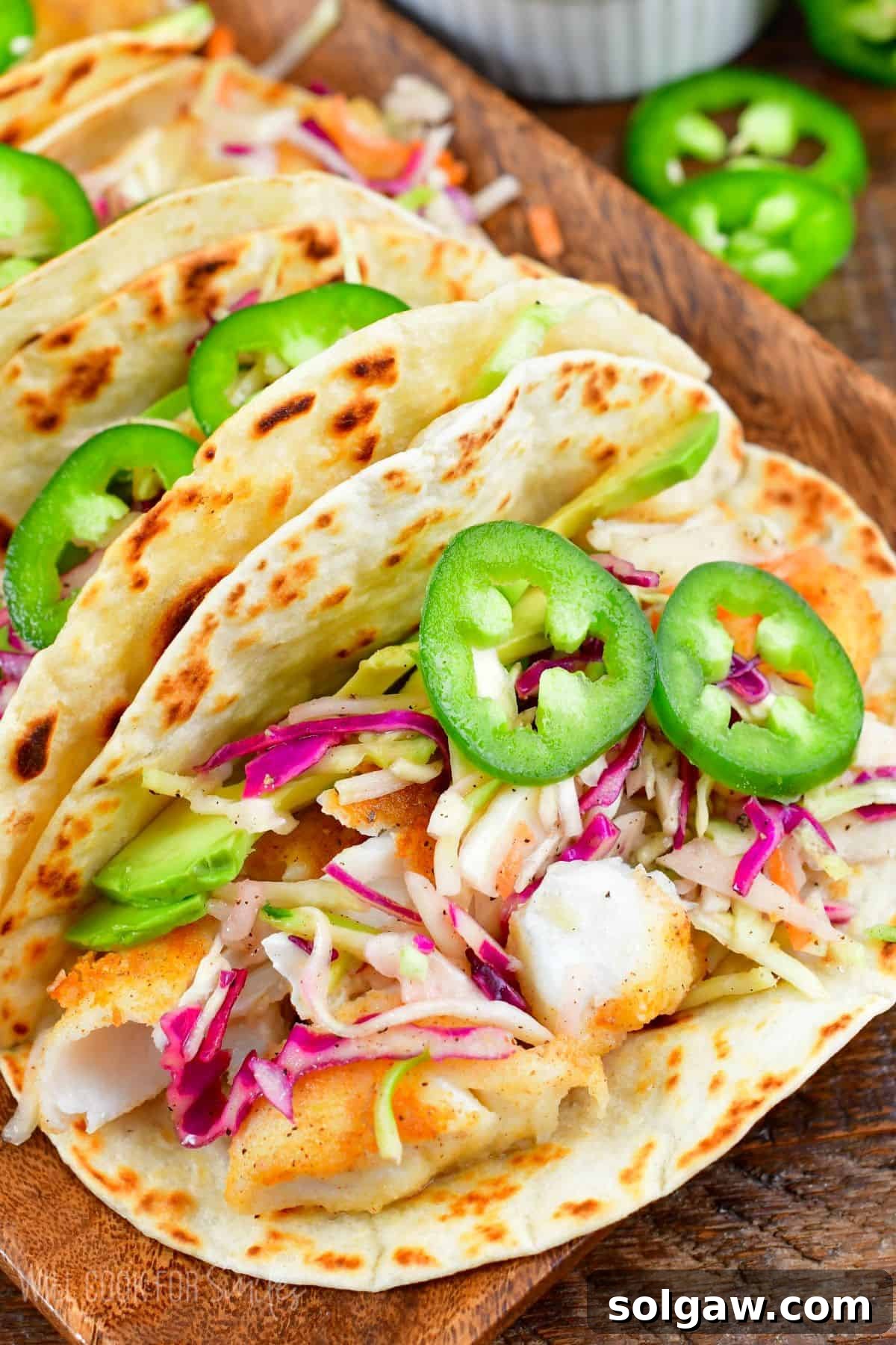 Fried fish tacos in flour tortilla with avocado, cabbage, jalapenos on top with a wood tray.