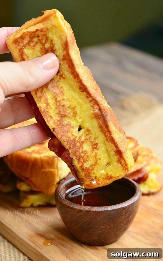 Close-up of a French Toast stick, dipped in glistening maple syrup, held ready to eat, highlighting its golden exterior and rich texture.