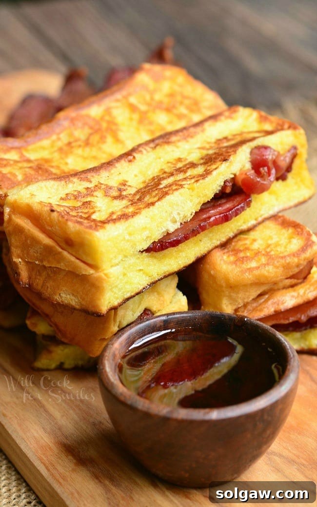 Close-up of golden brown Bacon Stuffed Brioche French Toast sticks stacked on a wooden cutting board with a small bowl of maple syrup for dipping.