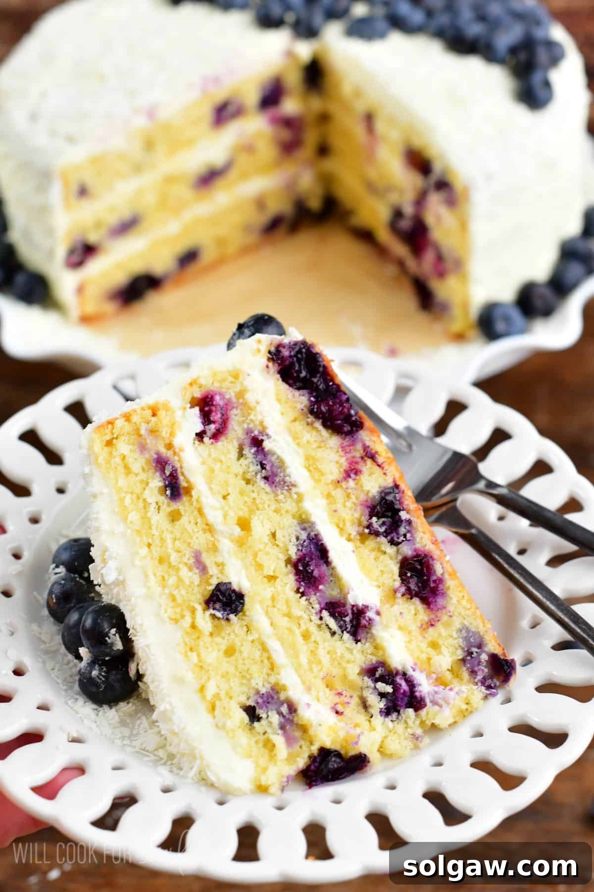 A beautifully sliced blueberry cake on a white plate, with the rest of the cake standing elegantly on a cake stand nearby. The cake features layers of fluffy yellow cake, rich cream cheese frosting, and vibrant fresh blueberries, highlighting its appeal as a perfect summer dessert.
