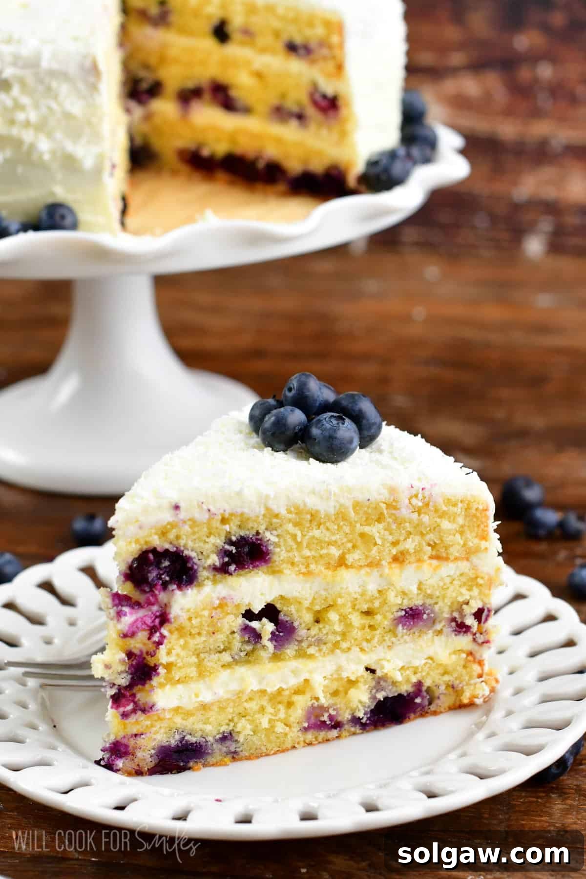 A generously frosted slice of blueberry cake sits on a plate, surrounded by fresh blueberries, with the remainder of the elegant layered cake visible in the background.