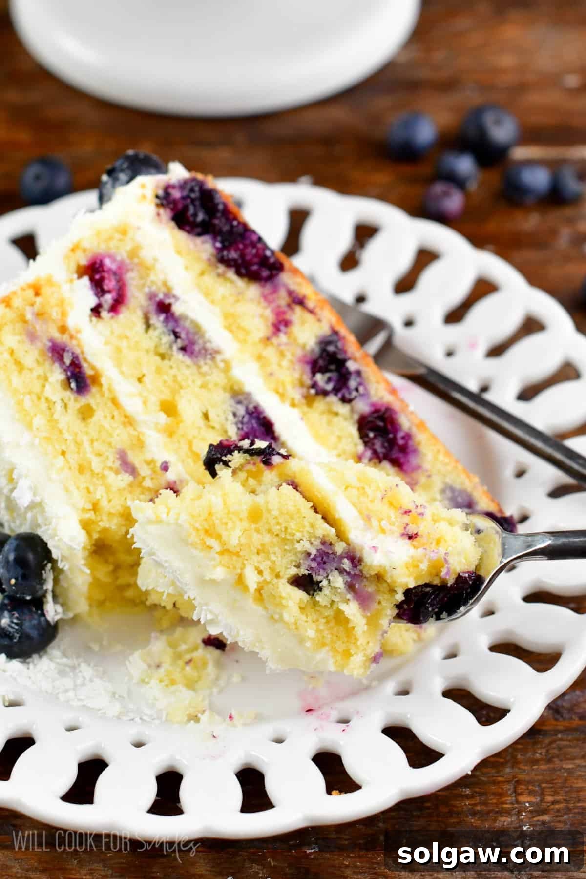 A fork lifts a tempting bite from a slice of blueberry cake, highlighting its moist texture and vibrant blueberries, all presented on a simple white plate.