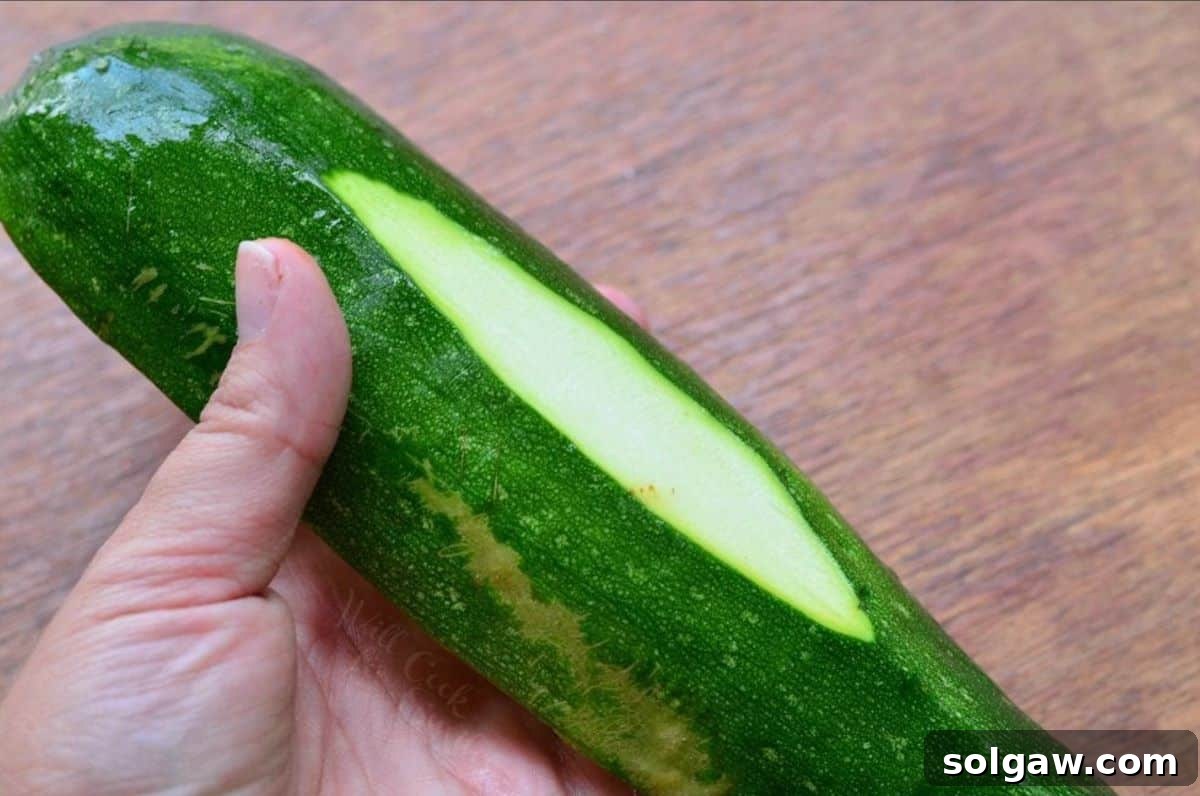 A hand holding a zucchini half, demonstrating how to slice a thin piece off the bottom for stabilization.