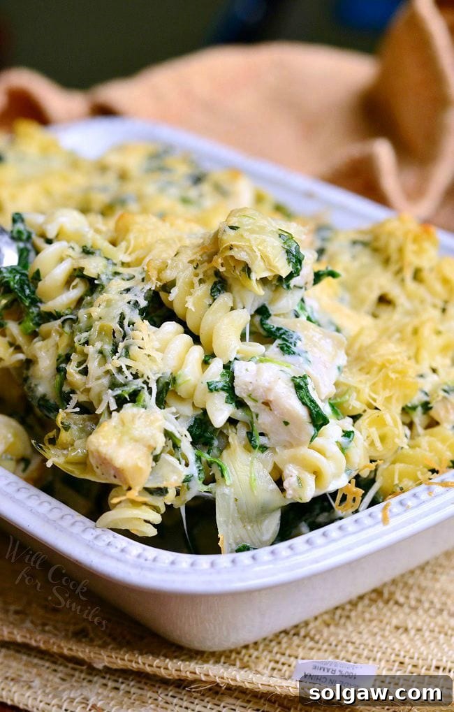 Spoon lifting up some of the Chicken Spinach and Artichoke Pasta that is served in a white casserole dish. 