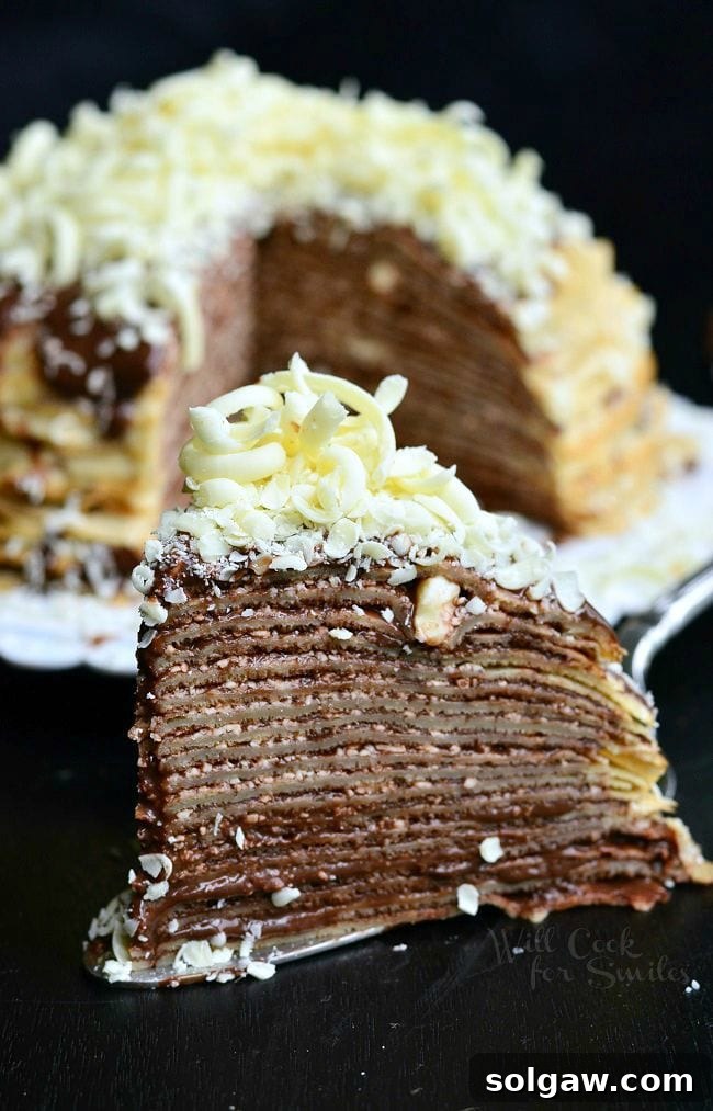 A slice of the Double Chocolate Chocolate Pudding Crepe Cake sits on a serving spatula. The stacked crepes make for thin layers. Chocolate is a key element of this cake. It's between all layers and then on top as well. There are also white chocolate shavings on top of this slice of cake. The rest of the cake fades into the background. 