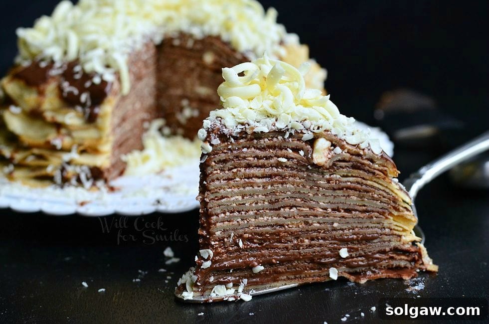 A slice of the Double Chocolate Chocolate Pudding Crepe Cake sits on a serving spatula. The stacked crepes make for thin layers. Chocolate is a key element of this cake. It's between all layers and then on top as well. There are also white chocolate shavings on top of this slice of cake. The rest of the cake fades into the background.