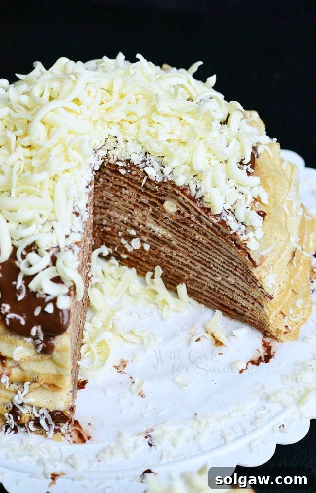 Double Chocolate Chocolate Pudding Crepe Cake is served on a white dish. The crepes are stacked on one another with filling between layers. The cake is topped with more chocolate pudding and white chocolate shavings. One piece of the cake is missing allowing the inside layers to be seen.