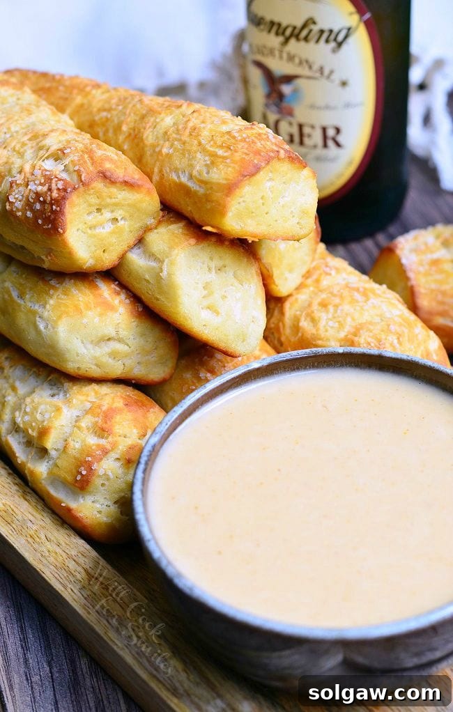 A mound of Homemade Soft Pretzels placed on a wooden board. Pretzels are a nicely golden brown. In front of the pretzels, is a bowl of beer cheese dip. In the background, there is a bottle of beer.
