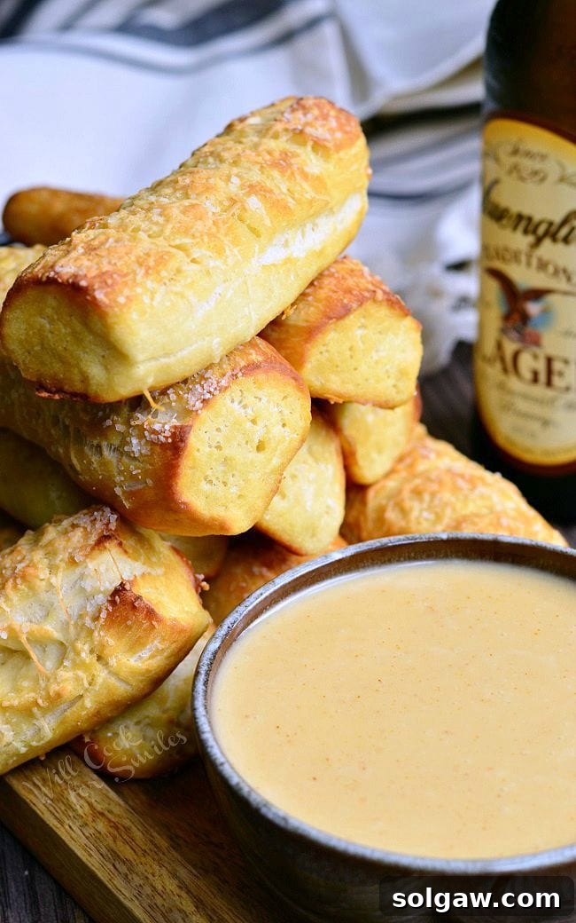 A mound of Homemade Soft Pretzels placed on a wooden board. Pretzels are a nicely golden brown. Infront of the pretzels, is a bowl of beer cheese dip. In the background, there is a bottle of beer.