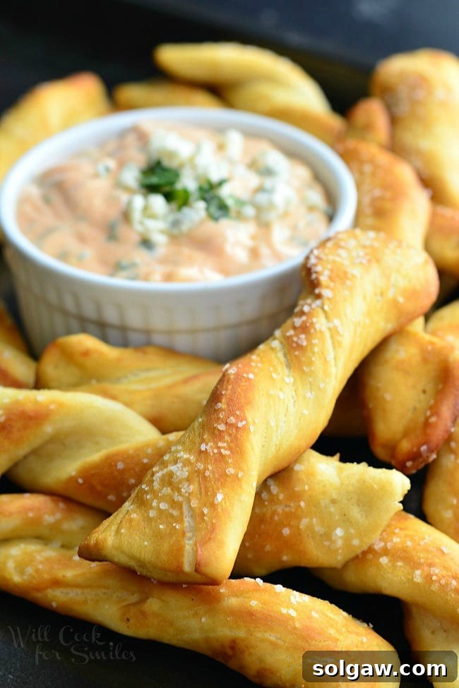 Homemade Soft Pretzel Twists served around a small, white dish containing Creamy Buffalo Sauce. Pretzels are a nice golden brown with coarse salt on top. The buffalo sauce is garnished with minced parsley and blue cheese crumbles.