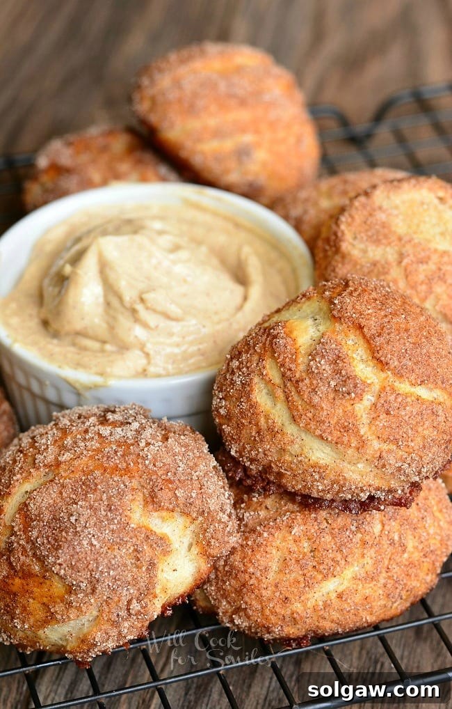 Snickerdoodle Pretzel Puffs served on a wire rack around a small, white dish containing a creamy cinnamon dip. These soft, fluffy little pretzel buns are made and coated with cinnamon and sugar.
