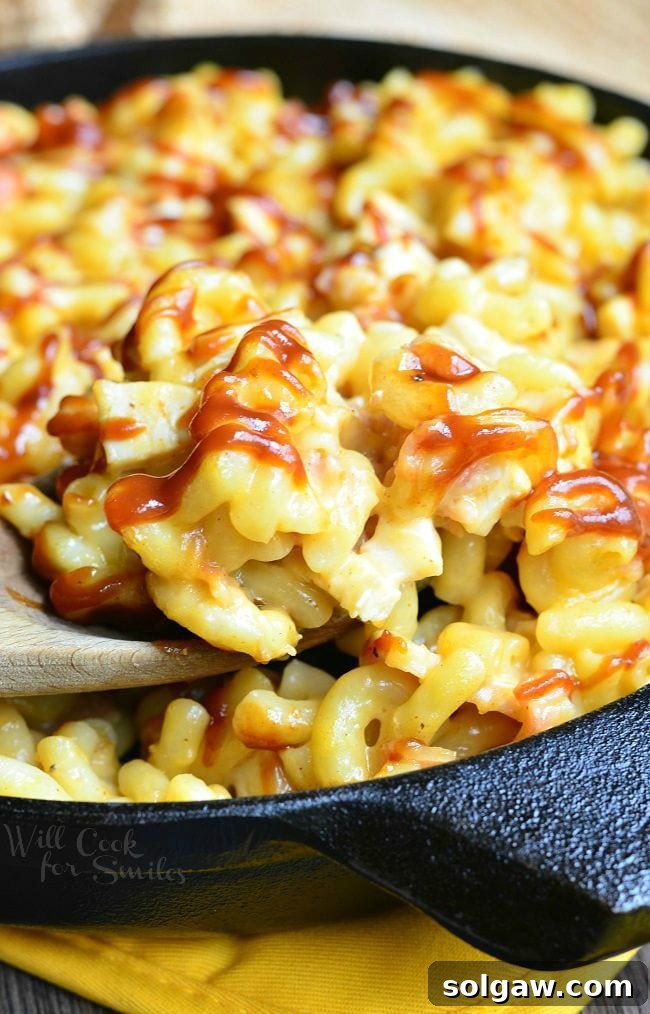 A wooden spoon is dipped into the skillet of Smokehouse Mac and Cheese. There are noodles, chicken, bacon and lots of cheese. The top of the Mac and Cheese is drizzled with BBQ sauce.