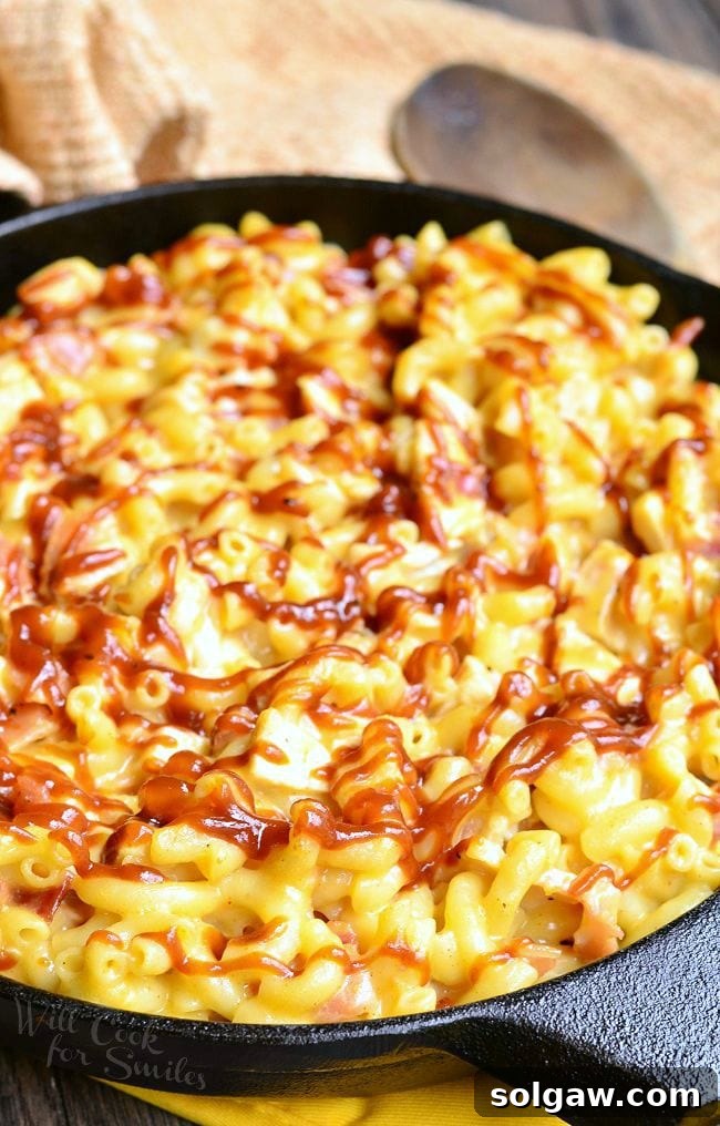 Presented is a black skillet full of Smokehouse Mac and Cheese. There are noodles, chicken, bacon and lots of cheese. The top of the Mac and Cheese is drizzled with BBQ sauce.