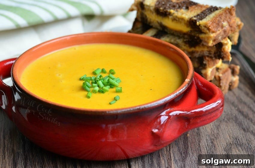 Red clay crock filled with pumpkin ale soup on a wooden table with a cut up grilled cheese in the background and a white and green cloth in the background to the left