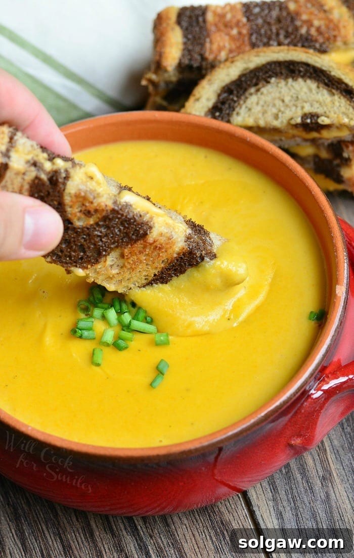 Dipping a grilled cheese sandwich into Pumpkin Ale Soup, garnished with chives in an orange soup bowl.