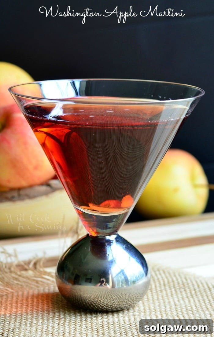 Side view of a decorative martini glass with Washington Apple Martini on a brown placemat with a bowl of apples in the background to the left