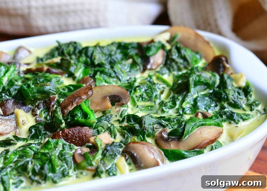 close up view of a white baking pan filled with creamed spinach and mushroom in white wine sauce on a wooden table with a white cloth behind