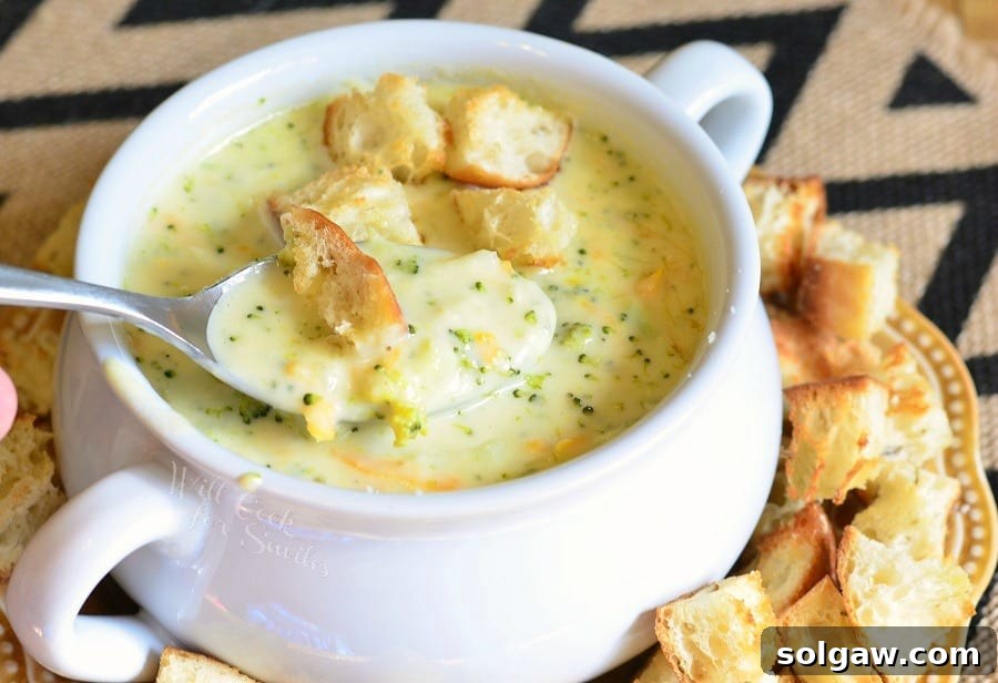 White crock filled with asiago cheese soup on a yellow plate with cut bread croutons scattered around the bowl placed on the plate viewed close up with a spoon holding one portion of soup