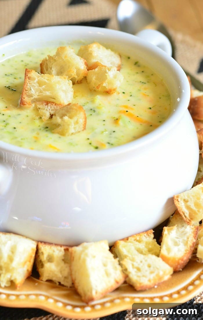 White crock filled with asiago cheese soup on a yellow plate with cut bread croutons scattered around the bowl placed on the plate with a silver spoon in the background