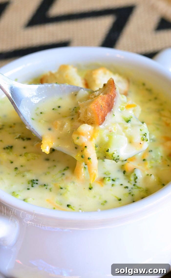 White crock filled with asiago cheese soup on a yellow plate with cut bread croutons scattered around the bowl placed on the plate with a spoon pulling out one portion of soup