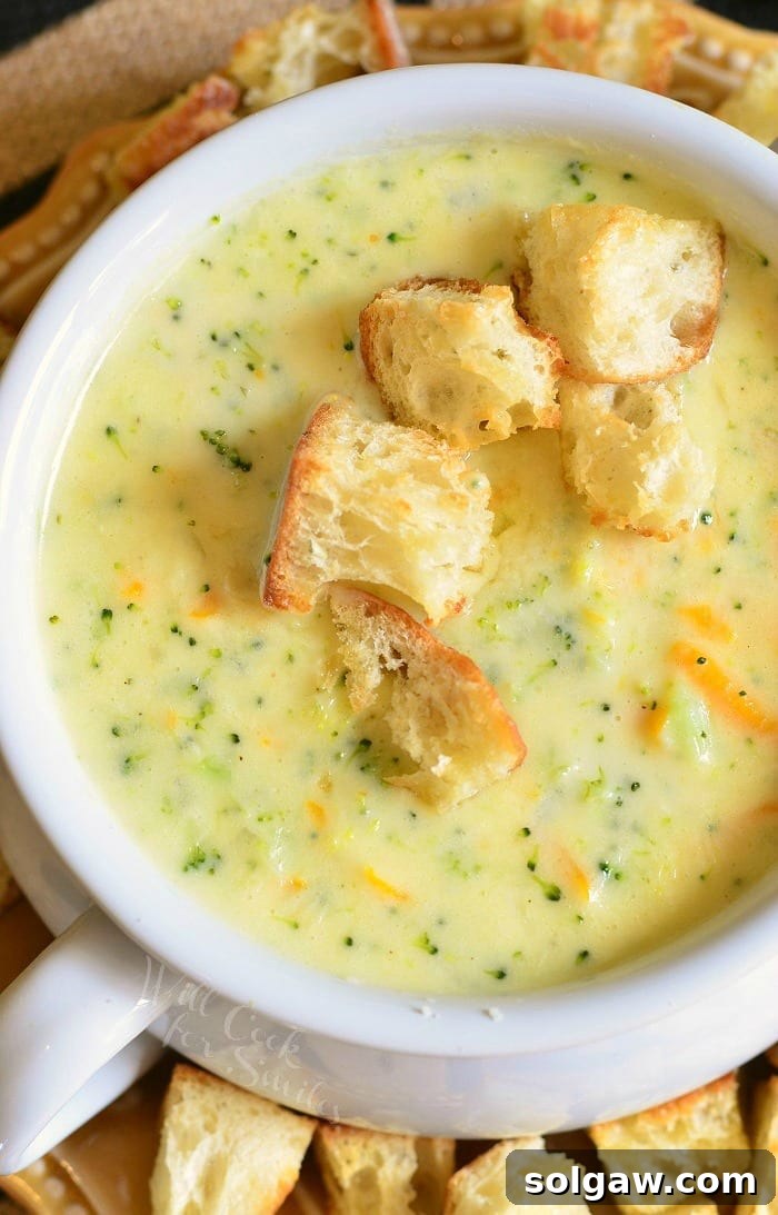 White crock filled with asiago broccoli cheese soup on a yellow plate with cut bread croutons scattered around the bowl placed on the plate as viewed from above