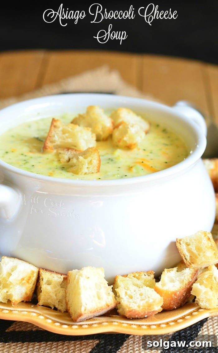 White crock filled with asiago cheese soup on a yellow plate with cut bread croutons scattered around the bowl placed on the plate on a black and white placemat on a wooden table