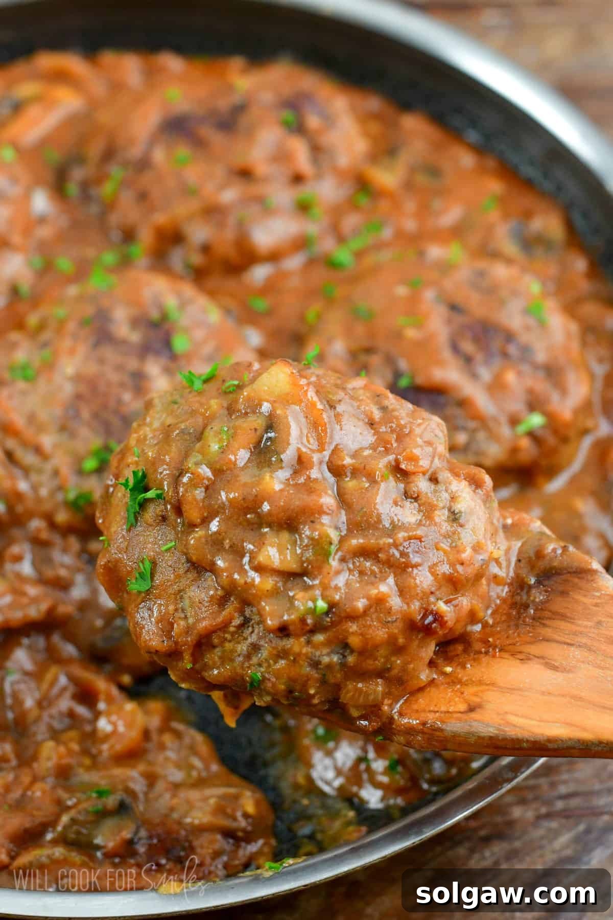 Holding a Salisbury steak on a wood spatula with the rest of the steaks in a pan.