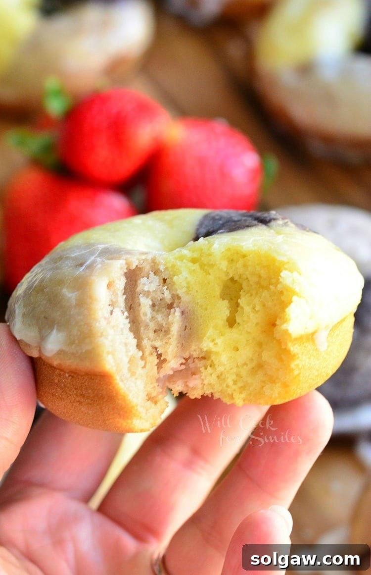 A hand holding a Neapolitan Glazed Doughnut with a bite taken out, revealing the soft, moist, multi-layered interior of chocolate, vanilla, and strawberry.