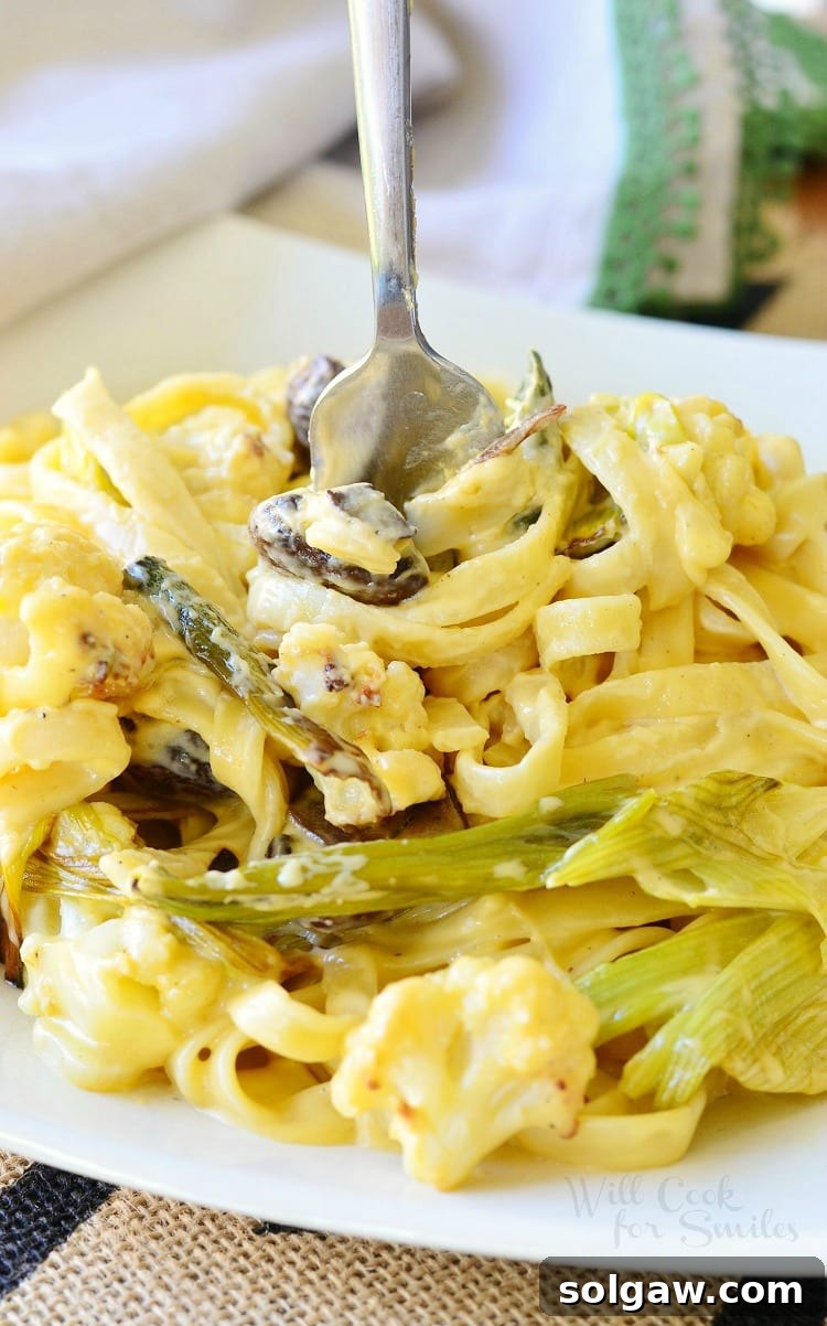 Close-up of a fork piercing into creamy Roasted Cauliflower, Leek, and Mushroom Fettuccine in Garlic Cheese Sauce, in a bowl.
