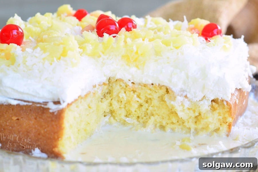 A Pina Colada Tres Leches Cake with a slice removed, revealing its moist layers, garnished with fluffy white coconut frosting, golden pineapple pieces, and bright red cherries.