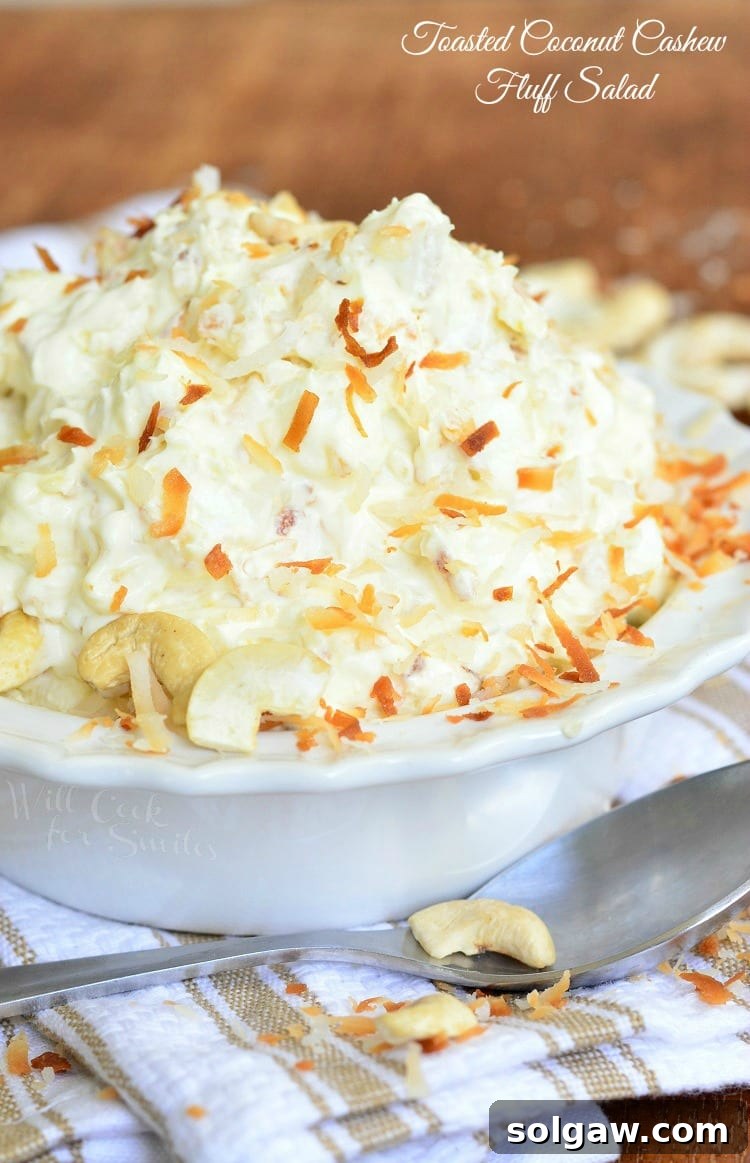 A beautifully presented Toasted Coconut Cashew Fluff Salad, topped with toasted coconut and cashews, in a pristine white bowl on a white and brown kitchen towel, with a spoon resting across the front, inviting a taste.