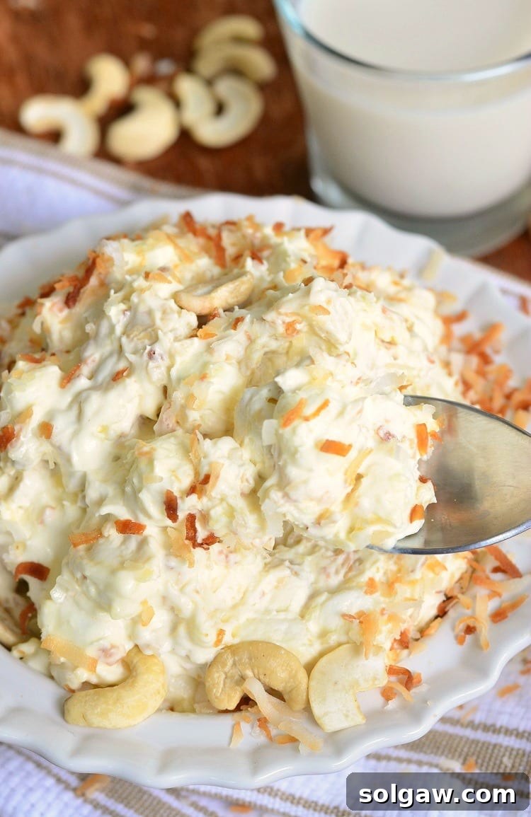 A top-down view of the Toasted Coconut Cashew Fluff Salad in a white bowl, resting on a white and brown kitchen towel, with a spoon scooping out a generous portion, showcasing its creamy texture and rich toppings.