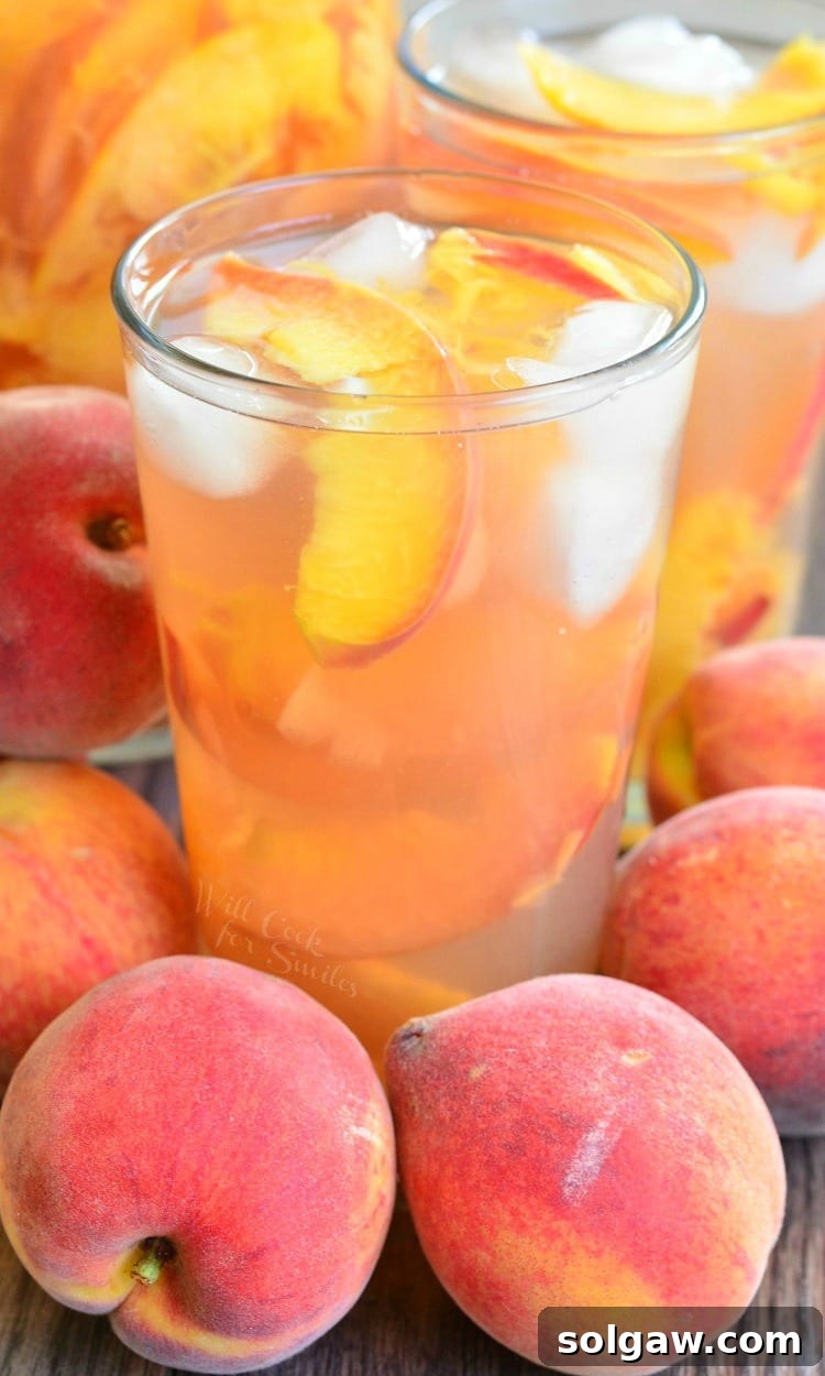 A tall glass of Ginger Peach and Honey Iced Green Tea, garnished with fresh peach slices and ice cubes, sits on a rustic wooden table. Around the glass are whole ripe peaches, suggesting the fresh ingredients used in the drink.