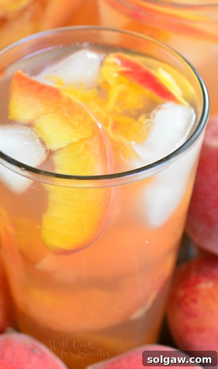 Close-up shot of Ginger Peach and Honey Iced Green Tea in a glass, showcasing the amber liquid, ice, and delicate peach slices. The surrounding table is adorned with more fresh peaches, emphasizing the key ingredient.