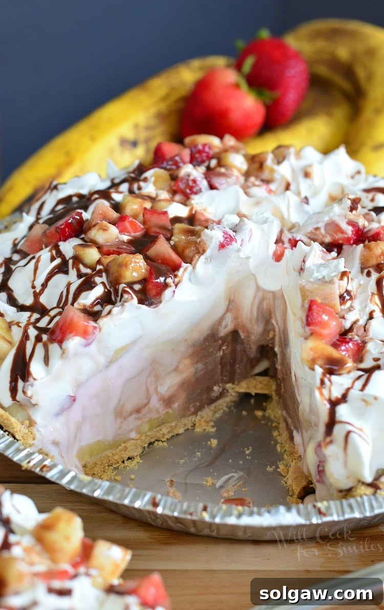 Banana Split Ice Cream Pie in a disposable metal pie pan with whipped cream, strawberries, banana, and chocolate syrup on top with a slice taken out
