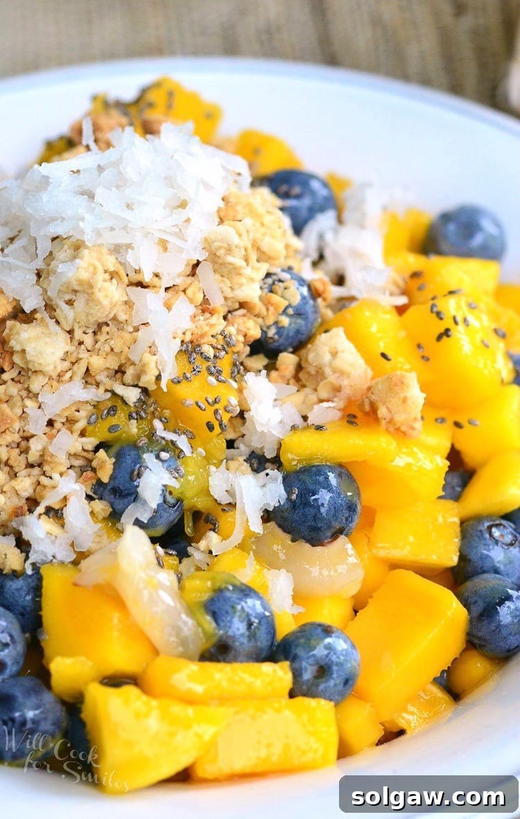 A vibrant bowl featuring chunks of ripe mango, peeled lychee fruit, and fresh blueberries, topped with golden granola and toasted coconut flakes.