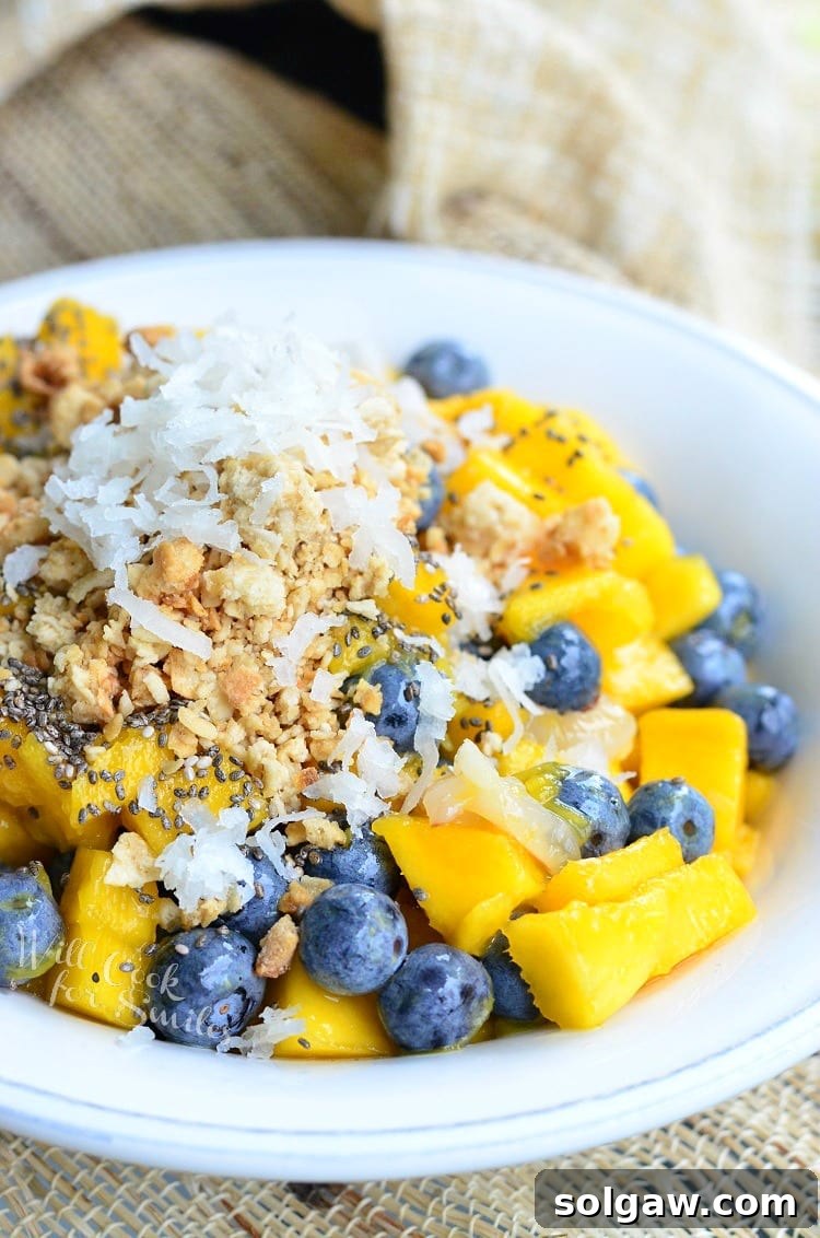 Close-up shot of a tropical fruit bowl with mango, lychee, and blueberries, generously topped with crunchy granola and sweet coconut flakes, ready to be enjoyed.
