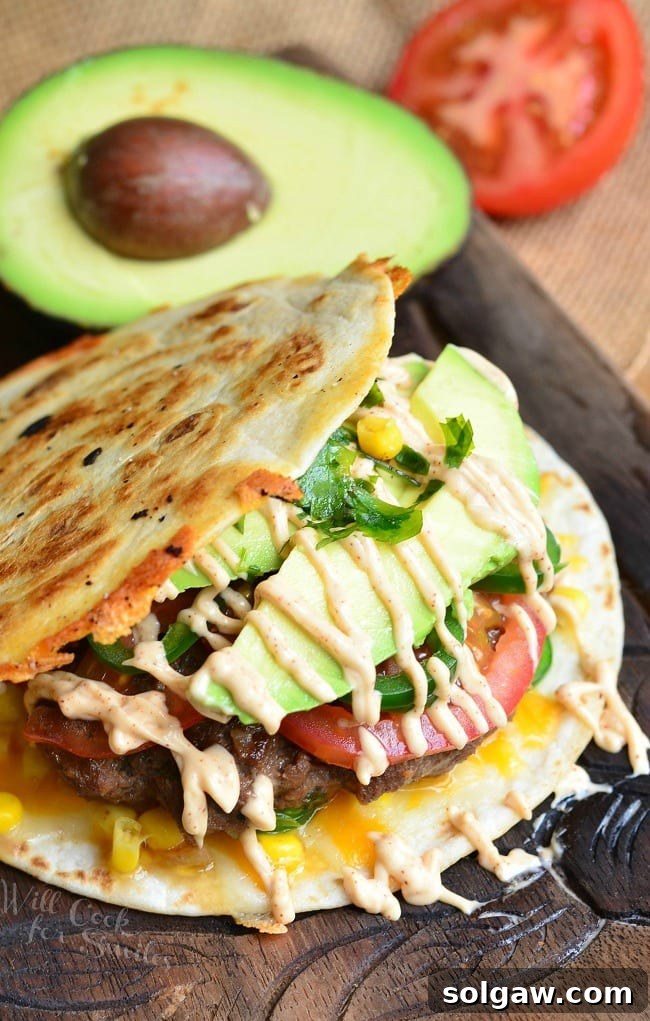 A unique burger sandwiched between tortillas with avocado, tomato, and homemade sauce, with fresh ingredients in the background
