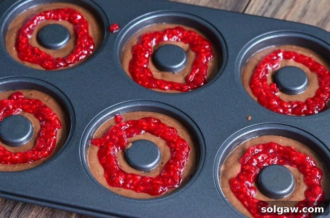 A close-up view of a doughnut baking pan, showing individual cavities perfectly filled with chocolate batter and a dollop of raspberry jam at the center, ready for baking.