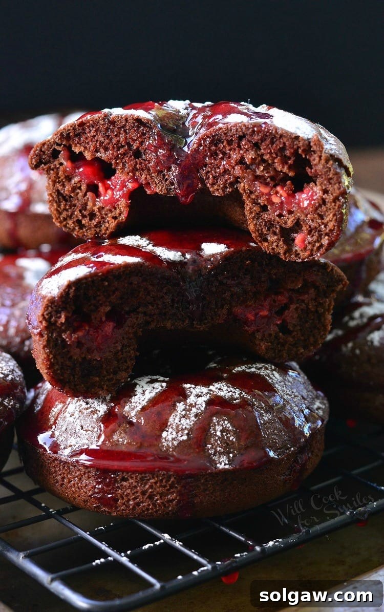 A stack of Raspberry Chocolate Doughnuts, with the top one enticingly cut in half to reveal its raspberry jam filling, all arranged on a cooling rack and dusted with powdered sugar and raspberry sauce.