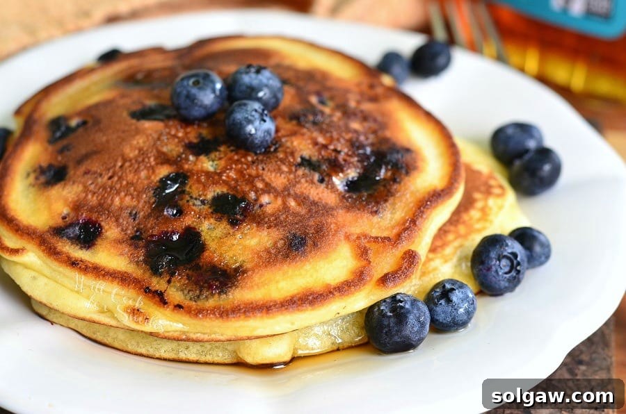 Single serving buttermilk pancakes topped with syrup and blueberries on a white plate with a fork in the background to the right as viewed close up